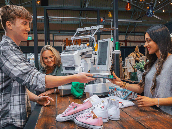 Drie mensen in interactie met de Brother PR1055X borduurmachine, met ingebouwde integratie met de BEaaS web app. Twee geborduurde schoenen worden vlakbij getoond.